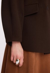 Brązowy wełniany żakiet z klapami i kieszenią, zestawiony z brązową zamszową spódnicą. Dłoń zdobiona złotym pierścionkiem z marmurowym kamieniem.