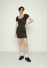 Mulher com cabelo escuro curto, usando óculos de sol âmbar, um vestido mini preto com estampado de flores amarelas, meias brancas e mocassins pretos, posando contra uma parede simples.