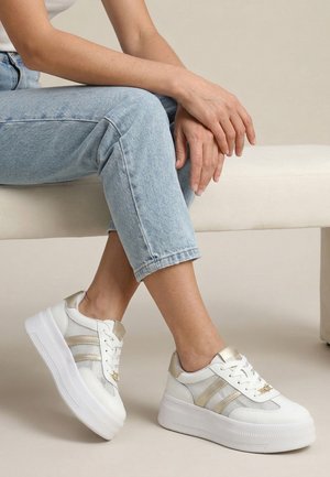 Osoba siedząca na jasnej ławce ubrana w jasnoniebieskie dżinsy i białe sneakersy na platformie ze złotymi paskami i detalami.