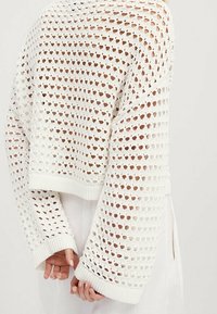 Valkoinen avoin neulepaita, jossa on lyhyt malli, pitkät hihat ja ribbaushelma. Teksturoitu kuvio, jossa on sydämen muotoisia aukkoja.