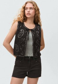 Zwart cropped vest met paisley borduursel en decoratieve kralen, gesloten met een strik. Draag het over een gestructureerde grijze top en gecombineerd met zwarte shorts.