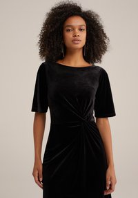 Black velvet dress with a boat neckline, short sleeves, and a twisted knot detail at the waist. Soft texture and fitted design.