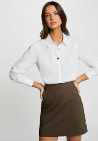 Camicia bianca con colletto classico e bottoni a forma di cuore, abbinata a una mini gonna fitted verde oliva con dettagli di bottoni laterali.