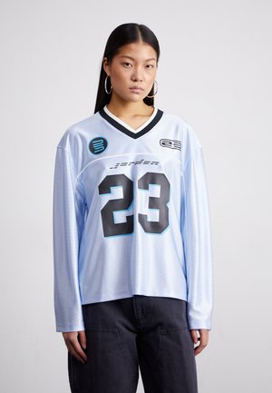 Light blue long-sleeve jersey featuring a black number "23," black V-neck collar, and logo patches in contrasting colors. Textured fabric with a relaxed fit.