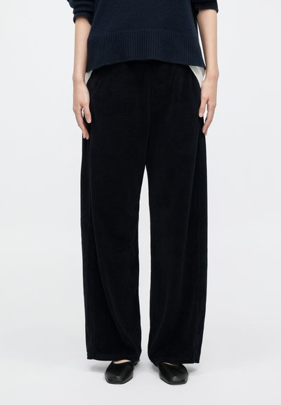 Άτομο που φορά λινά μαύρα παντελόνια, μαύρα παπούτσια και σκουρόχρωμο πουλόβερ, στέκεται μπροστά σε ένα απλό φωτεινό φόντο.