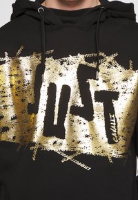 Černá mikina s kapucí s velkým, opotřebovaným zlatým nápisem "JUST" přes přední stranu, s propracovanými vzory a texturovaným povrchem.