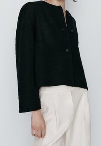 Giacca nera corta con una trama testurizzata, scollo rotondo e chiusure a bottone. Presenta tasche frontali e maniche lunghe. Si abbina a pantaloni di colore chiaro.