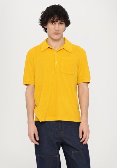 Gul polo skjorte med teksturert stoff, korte ermer, krage, tre knapper og en brystlomme. Bært med mørkeblå denimbukser.