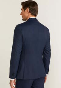 Mörkblå kavaj med texturerat tyg, strukturerad passform, enkel vent, och svarta knappdetaljer på ärmarna.