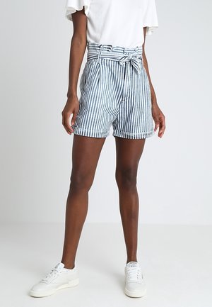 Kvinde iført blå og hvide lodrette stribede shorts med højtaljet bælte, hvid top og hvide sneakers, stående på en ensfarvet baggrund.