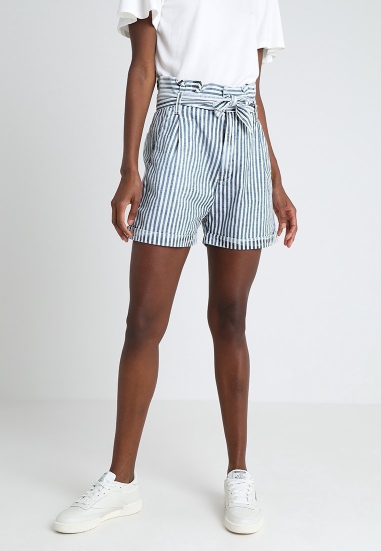 Femme portant un short à rayures verticales bleu et blanc taille haute avec une ceinture nouée, un haut blanc et des baskets blanches, debout sur un fond uni.