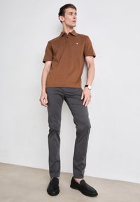 Barna póló ing két gombbal, szürke, slim fit nadrág, és fekete slip-on cipők. Minimalista dizájn, sima textura, és diszkrét márkajelzés az ingén.