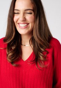 Rode gebreide trui met een V-halsontwerp, voorzien van een verticale ribbelstructuur. Een gouden smalle chokerhalsketting versterkt de simpele, nette uitstraling.