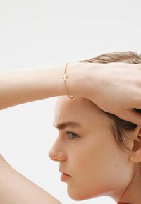 Brazalete de oro con un pequeño colgante de cruz, diseñado con una delicada cadena. La textura es suave y el color es un oro amarillo pulido.