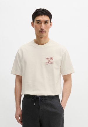 Mężczyzna noszący kremową koszulkę z czerwonym koktajlem i napisem „Marc O'Polo” na piersi, w połączeniu z czarnymi spodniami ze sznurkiem.