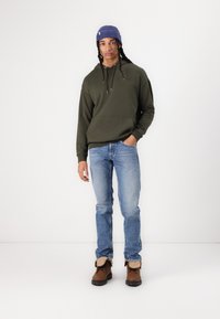 Olive green hoodie with a front pocket, blue ribbed beanie, light blue jeans, and brown boots with fur lining.