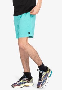 Shorts turquoise en coton, coupe décontractée, avec des poches latérales. Portés avec des chaussettes noires et des chaussures de sport multicolores, au design épuré.