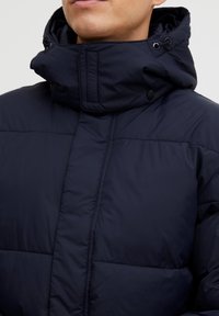 Marineblå puffjakke med høy krage, trykknapper og justerbare snorer. Glatt tekstur med quiltede seksjoner.