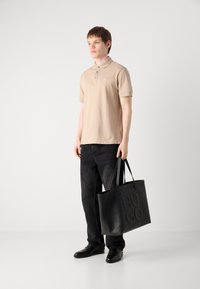 Bežna polo majica s gumbi, črne kavbojke in črna usnjena torba z embosiranim napisom "HUGO." Model nosi črne čevlje in stoji pred nevtralno ozadje.