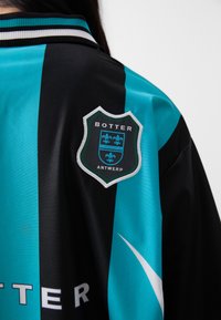 Fekete és türkiz sportdzseki bordázott gallérral, hímzett pajzslogóval, amelyen a "BOTTER" és az "ANTWERP" szöveg szerepel.