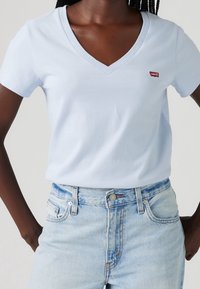 Světle modré tričko s výstřihem do V vyrobené z bavlny, s krátkými rukávy, se malým červeným logem Levi's na hrudi, v kombinaci se světle modrými džíny.