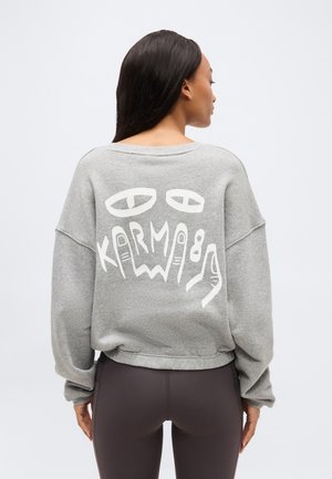 Femme de dos portant un sweat-shirt gris avec des yeux graphiques blancs et les lettres formant le mot « Karma » au dos, assorti à des leggings foncés.