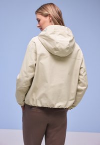 Donna di spalle che indossa una giacca beige con cappuccio e pantaloni marroni su uno sfondo azzurro chiaro.