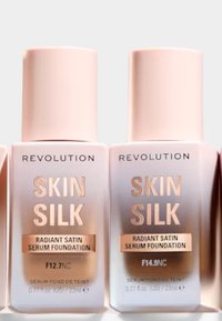 To frostat glassflasker med Revolution Skin Silk Radiant Satin Serum Foundation; lyserosa hetter, rosegull tekst, merket F12.7NC og F14.9NC.