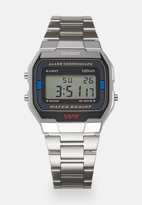 Montre numérique Casio avec un bracelet métallique en argent. Caractéristiques d'un cadran carré noir, d'un écran LCD et d'une étiquette de résistance à l'eau en rouge.