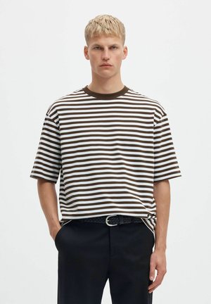 Un hombre luce una camiseta blanca holgada con rayas horizontales de color marrón oscuro, pantalones oscuros y un cinturón trenzado, sobre un fondo liso y claro.