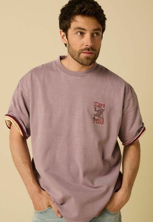 Homme aux cheveux bouclés foncés et à la barbe, portant un t-shirt ample mauve avec des manches retroussées et un petit motif rouge sur la poitrine, debout les mains dans les poches.
