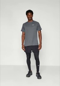 Sivý krátky rukáv Nike športový dres, čierne šortky a čierne kompresné legíny, s čiernymi športovými topánkami. Pozadie je jednoduché a svetlé.