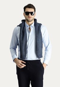 Camicetta blu chiaro con bottoni, maglione a coste blu navy appoggiato sulle spalle, occhiali da sole neri e pantaloni blu navy. Abbigliamento casual con dettagli testurizzati.