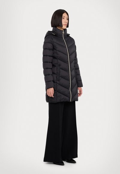 Manteau doudoune noir matelassé avec capuche, présentant une fermeture éclair dorée, un motif en chevrons et des manches longues. Associé à un pantalon noir à jambes larges.