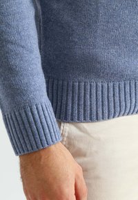 Close-up van een hand en pols die een blauwe gebreide trui met geribbelde manchetten draagt over beige broek.