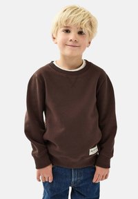 Ruskea huppari, jossa on ribbineck ja hihansuut, ja siinä on pieni logo-patch helmassa. Päällä valkoinen T-paita. Näkyvissä siniset farkut.