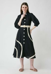 Schwarzes Hemdkleid mit Puffärmeln, cremefarbenen Akzenten und einem Stoffgürtel mit einer runden Schnalle. Modell kombiniert mit braunen Sandalen.