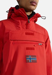 Rød lett jakke med høy krage, justerbar hette, lomme med glidelås foran, og et merke med det norske flagget. Glatt tekstur, funksjonell design.