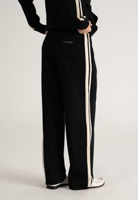 Person wearing black wide-leg pants with white side stripes and a back pocket, paired with white shoes on a neutral background.