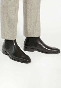 Black leather Chelsea boots with a smooth finish, elastic side panels, and a slight heel, paired with gray tailored trousers.