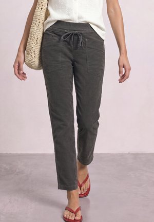 Personne portant un pantalon gris foncé à cordon, un haut blanc sans manches, des sandales rouges, portant un sac à épaule en crochet beige, marchant sur un sol gris clair.
