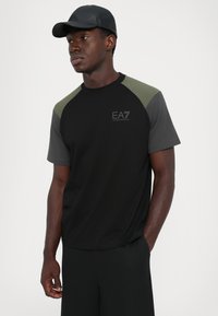 Young man wearing black cap and black and gray EA7 Emporio Armani t-shirt, standing with one hand in pocket against plain background.