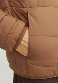Brūnā pildīta jakas ar vērpuma tekstūru. Iespējas ir ribota aproce un redzamais šuvums. Materiāls izskatās viegls un gluds.