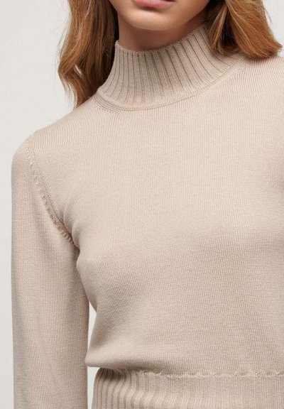 Beige neulepusero, jossa on ribatty poolokaulus, pitkät hihat ja istuva vyötärö. Kankaan pinta on pehmeä ja tekstuuri on elävä.