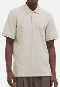 Jasno beżowa koszulka polo z teksturowanej tkaniny, z krótkimi rękawami, tradycyjnym kołnierzykiem i zapięciem na trzy guziki. Minimalistyczny design.