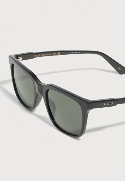 Lunettes de soleil Gucci à monture carrée noire avec verres teintés foncés, branches griffées et inscription "Made in Italy" à l'intérieur des branches.