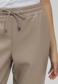 Beige neplederen joggers met een elastische tailleband en verstelbare trekkoord. Voorzien van glanzende metalen uiteinden op het trekkoord. Glad oppervlak.