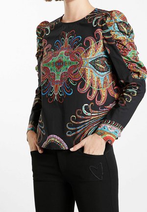Woman wearing a black blouse with colorful paisley patterns and puffed sleeves, paired with black pants featuring a stitched heart on the pocket.