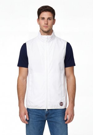 Man draagt een mouwloos wit vest met rits over een marineblauw T-shirt met korte mouwen en blauwe spijkerbroek, staand tegen een effen witte achtergrond.