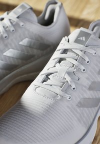 Scarpe atletiche bianche con tessuto testurizzato, accenti argento e lacci piatti. Presentano tre strisce grigie sul lato e una suola imbottita.